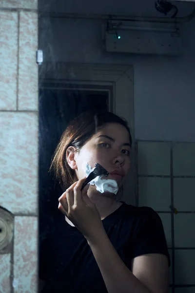 女人手里拿着剃须刀 站在浴室的镜子前 把剃须泡沫涂在下巴上 免版税图库照片
