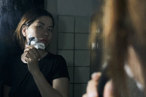 女人手里拿着剃须刀 站在浴室的镜子前 把剃须泡沫涂在下巴上 图库图片