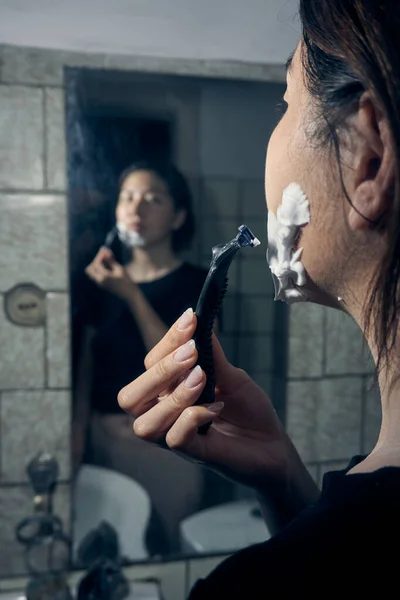 女性は手にカミソリを持ちバスルームミラーに立っている間に顎にシェービングフォームを適用します ロイヤリティフリーのストック画像