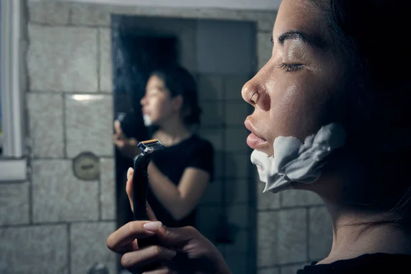 女人手里拿着剃须刀 站在浴室的镜子前 把剃须泡沫涂在下巴上 图库照片