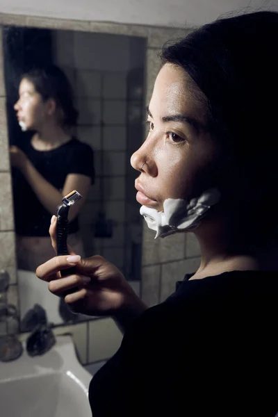 女人手里拿着剃须刀 站在浴室的镜子前 把剃须泡沫涂在下巴上 图库照片