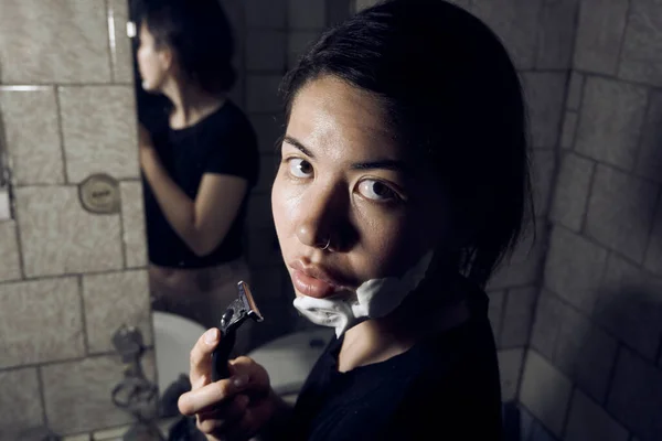 女人手里拿着剃须刀 站在浴室的镜子前 把剃须泡沫涂在下巴上 免版税图库照片