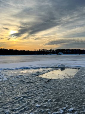 Güneş karla kaplı bir plajın ve donmuş bir göletin üzerinde batıyor. Kıyıya çekilen bir tekne görülüyor..