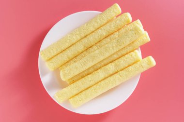 Corn crispy sticks or flip snacks on a plate on pink background, top view