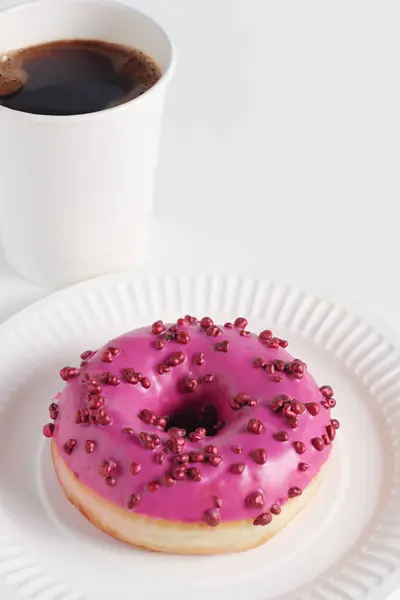 Pink donut with red sugar balls sprinkles and paper glass of coffee on a white background