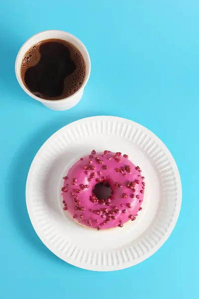 Pink donut with red sugar balls sprinkles and paper glass of coffee on blue background, top view. Disposable eco-friendly tableware