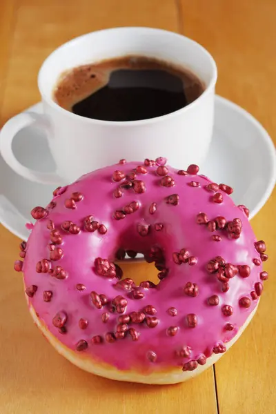 Pink donut with red sugar balls sprinkles and cup of coffee on wooden table close-up