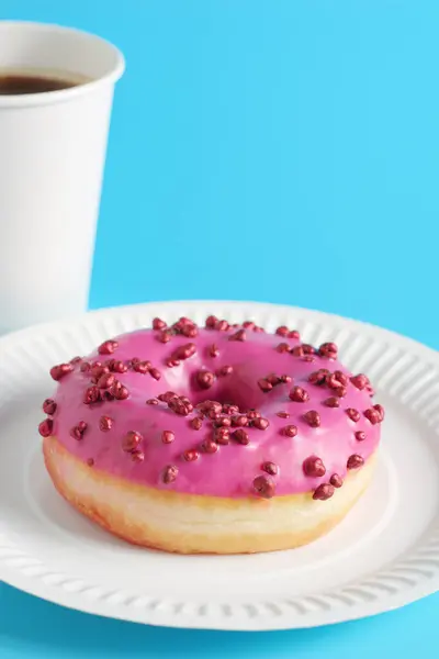Pink donut with red sugar balls sprinkles and paper glass of coffee on blue background. Space for text