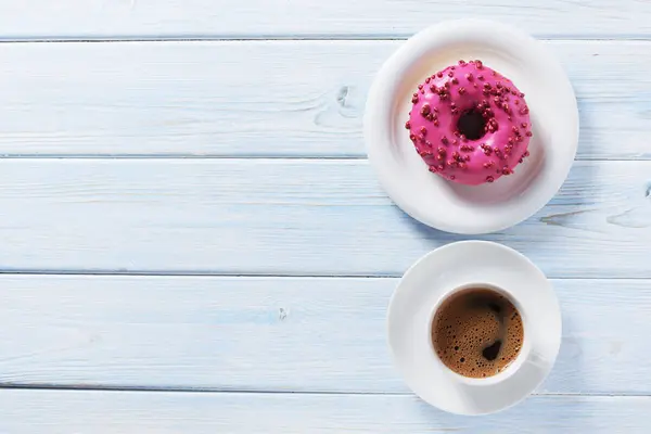 Pink donut with red sugar balls sprinkles and cup of coffee on blue wooden table, top view with space for text