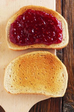 İki dilim kızarmış beyaz ekmek, biri reçelli, diğeri reçelsiz, kesme tahtasının üzerinde, eski ahşap arka planda, üst tarafta.