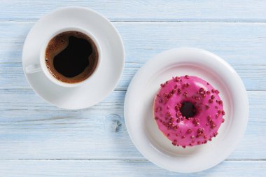Kırmızı şekerli pembe çörek, serpiştirmeler ve mavi ahşap masada bir fincan kahve, üst manzara.