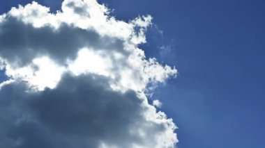 Sun in a blue sky. White and gray clouds drift across the image, obscuring the sun. Beautiful cottony cumulus clouds in timelapse. Sunbeams appear between and through the clouds.