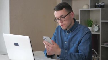 Gözlüklü genç yakışıklı bir adam, telefonun içeriğine bakıyor, duygusal olarak el kol hareketi yapıyor. Sıcak bir ev ortamı. Yavaş çekim...