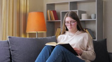 Uzun kahverengi saçlı, gözlüklü güzel bir kız öğrenci kanepede otururken bir kitap okur. Ev ortamı. Yavaş çekim..