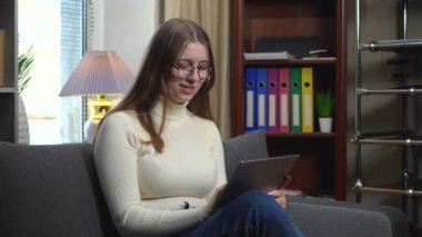 Uzun kahverengi saçlı güzel bir kız, bir tabletle iletişim kuruyor, mesaj atıyor ve e-posta atıyor, gülümsüyor, sıcak, sade bir ortamda kanepede oturuyor. Yavaş çekim.