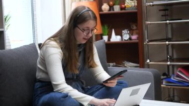 Uzun sarı saçlı, kulaklıklı genç, güzel bir kız, bilgisayardan mesaj atar, telefondan veri okur, kanepede oturur, ev ortamında. Yavaş çekim