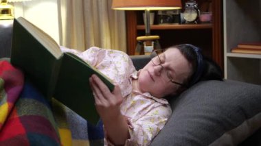 Yaşlı bir kadın, gözlük takıyor, kanepede yatıyor, battaniyeyle örtülmüş, yatmadan önce kitap okuyor. Yavaş çekim.
