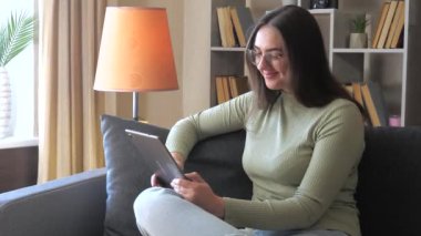 Genç ve güzel bir kız, gözlük takıyor, evdeki kanepede oturuyor, tablet kullanan bir arkadaşıyla iletişim kuruyor, gülümseyerek selamlıyor ve el sallıyor. Yavaş çekim.