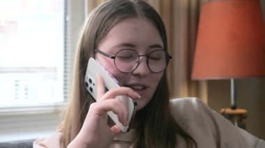 Gözlüklü genç bir kız, ev ortamında, kanepede oturuyor, ailesiyle telefonda konuşuyor, ilişkileri hakkında konuşuyor ve gülümsüyor. Yavaş çekim.