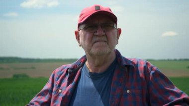  Ekose gömlekli ve kırmızı beysbol şapkalı yaşlı bir çiftçi bir tarlada duruyor ve mavi gökyüzüne karşı manzarayı inceliyor. Yavaş çekim.