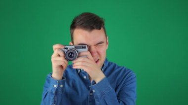 Mavi gömlekli genç yakışıklı bir adam eski bir kamerayla fotoğraf çekiyor, elinde tutuyor. Yeşil arka plan. Yavaş çekim.