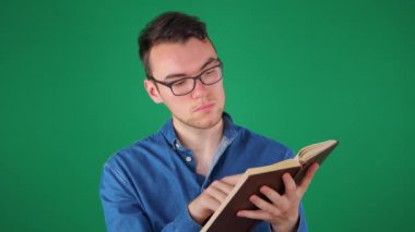 Gözlüklü ve mavi gömlekli genç bir adam kitap okuyor. Yeşil arka plan..
