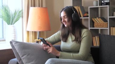 Genç, güzel bir kız öğrenci, kanepede oturuyor, telefonda müzik dinliyor, gülümsüyor. Sıcak ev ortamı