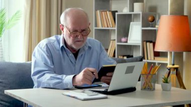 Yaşlı, gri saçlı bir iş adamı, mavi gömlekli, evde mali bir raporu kontrol ediyor, hesap makinesi ve bilgisayardaki verileri kontrol ediyor, ve bunları belgelere kaydediyor..