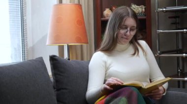 Gözlüklü genç bir kız, evdeki kanepede oturuyor, sıcak bir battaniyeyle örtülmüş, kurgu okuyor.