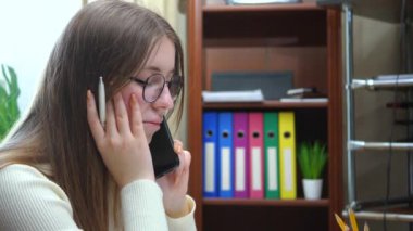 Genç, güzel bir kız menajer, gözlüklü, masasında oturan, elinde kalemle telefonla konuşan..