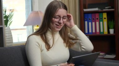 Genç ve güzel bir kız, gözlüklü beyaz bir bluz giyiyor, rahat bir ev ortamında kanepede oturuyor, tablet kullanarak internetten iletişim kuruyor, saçını düzleştiriyor ve gülümsüyor. Yavaş çekim.