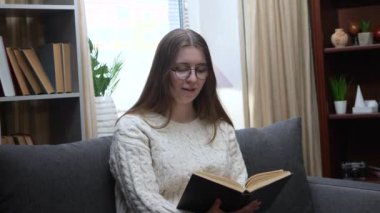 Genç, güzel bir kız, gözlüklü beyaz bir bluz içinde, rahat bir ev ortamında kanepede oturuyor, bir kitap okuyor. Yavaş çekim.