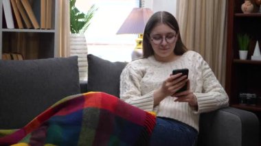 Genç ve güzel bir kız, gözlüklü beyaz bir bluz içinde, sıcak bir battaniyeyle kaplı, rahat bir ev ortamında kanepede oturuyor, bir SMS mesajı yazıyor ve gülümsüyor. Yavaş çekim.
