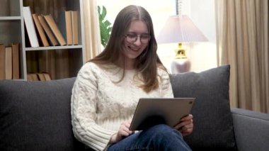 Genç ve güzel bir kız, gözlüklü beyaz bir bluz giyiyor, rahat bir ev ortamında kanepede oturuyor, tablet kullanarak annesiyle internetten iletişim kuruyor ve gülümsüyor. Yavaş çekim.