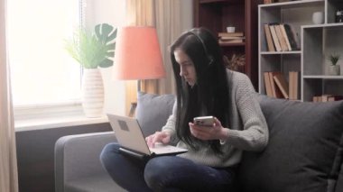 Koyu renk saçlı, gri bluzlu genç bir kız öğrenci kanepede oturuyor ve telefonundan bir bilgisayara veri aktarıyor. Mesafe öğrenme. Yavaş Hareket