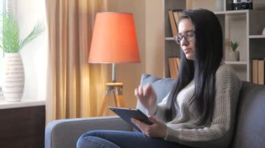 Koyu renk saçlı, gri bluzlu genç bir kız öğrenci kanepede oturuyor ve telefonundan bir bilgisayara veri aktarıyor. Mesafe öğrenme. Yavaş Hareket