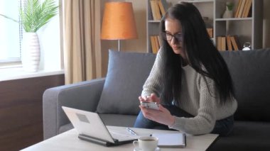 Koyu renk saçlı, gri bluzlu genç bir kız öğrenci kanepede oturuyor ve telefonundan bir bilgisayara veri aktarıyor. Mesafe öğrenme. Yavaş Hareket