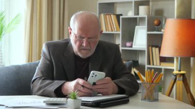 Ceketli yaşlı bir adam yıllık muhasebe raporu üzerinde çalışıyor, hesap makinesindeki verileri kontrol ediyor ve bilgisayarda birleştiriyor, bir belgeye yazıyor. Evden çalışıyorum. Yavaş çekim.