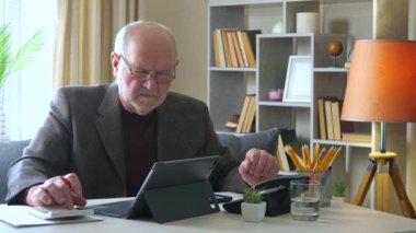  Ceketli yaşlı bir adam yıllık muhasebe raporu üzerinde çalışıyor, hesap makinesindeki verileri kontrol ediyor ve bilgisayarda birleştiriyor, bir belgeye yazıyor. Evden çalışıyorum. Yavaş çekim.