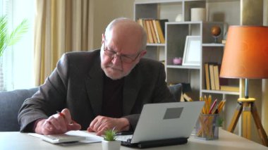  Ceketli yaşlı bir adam yıllık muhasebe raporu üzerinde çalışıyor, hesap makinesindeki verileri kontrol ediyor ve bilgisayarda birleştiriyor, bir belgeye yazıyor. Evden çalışıyorum. Yavaş çekim.
