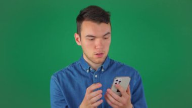 Mavi gömlekli genç bir adam telefonda konuşur, el kol hareketi yapar ve duygusal olarak gülümser. Yeşil arka plan. Yavaş çekim.