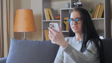 Genç ve güzel bir kız, dalgalı siyah saçlı, evdeki kanepede oturuyor, annesiyle telefonda konuşuyor, gülümsüyor. Rahat bir ev ortamı.