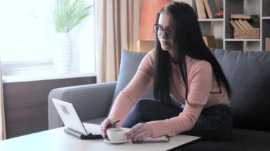  Gözlüklü ve pembe ceketli genç bir kız, bir öğrenci, kanepede oturuyor, bilgisayarla uğraşıyor. Kız çalışmayı bırakıyor, bir fincan sıcak kahve alıyor ve bir yudum alıyor. Rahat bir ev ortamı.