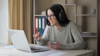 Gözlüklü ve kulaklıklı genç bir kız, bir öğrenci, arkadaşlarıyla telefon ve bilgisayar kullanarak iletişim kuruyor, duygusal olarak hareket ediyor ve ekranda gülümsüyor. Yavaş Hareket