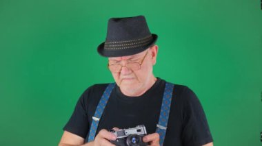 Şapkalı ve siyah tişörtlü yaşlı bir adam hatıra olarak fotoğraf çekmek için eski bir kamerayla fotoğraf çekiyor. Ağır çekim. Yeşil arkaplan