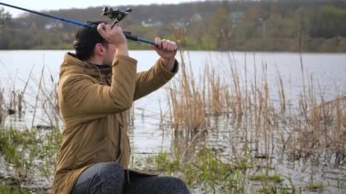  Mavi ceketli, gözlük takan genç bir adam, nehrin kenarında balık yakalama umuduyla bir olta atıyor, sessizliğin tadını çıkarıyor ve iyi bir avın hayalini kuruyor. Yavaş çekim.