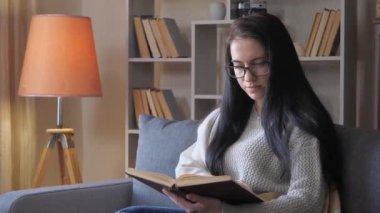  Genç bir kız öğrenci, esmer, gözlüklü ve gri kazaklı, roman okuyor, yavaşça kitabın sayfalarını çeviriyor. Yavaş çekim.