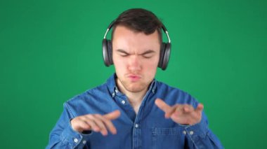  Mavi kot gömlekli genç bir adam kulaklıkla müzik dinler, ritme göre sallanır, gülümser ve dans eder. Yeşil arka plan. Yavaş çekim.