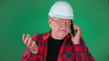 Yaşlı bir adam, müteahhit, beyaz koruyucu kasklı, gözlüklü, telefonla konuşan, el kol hareketi yapan. İş görüşmesi. Pembe arka plan. Yavaş çekim.