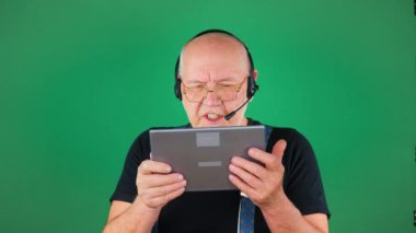 Gözlüklü, siyah tişörtlü ve pantolon askısı takan yaşlı bir adam tablet, jestler ve gülümsemeler kullanarak arkadaşlarıyla duygusal olarak iletişim kuruyor. Yeşil arka plan. Yavaş çekim.